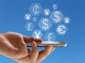 Mão segurando smartphone com ícones de diferentes moedas flutuando sobre fundo azul, representando bancos digitais com conta multimoeda.