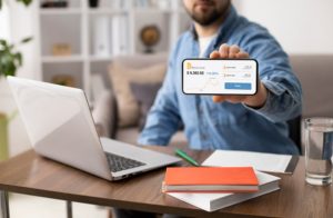 Homem sentado à mesa usando notebook e exibindo no celular a tela de acompanhamento de investimentos, simbolizando o uso de plataformas de empréstimo para gestão financeira.