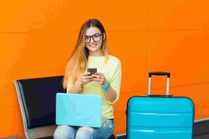 Mulher jovem sentada em um saguão com mala azul e laptop no colo, sorrindo enquanto usa o celular, representando a praticidade das contas digitais para viagens nacionais ao facilitar pagamentos e o controle financeiro durante deslocamentos.