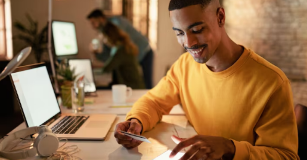 O Avanço do Microcrédito Digital no Brasil: Inclusão Financeira ou Endividamento em Massa? 1 Homem sorridente usando o celular e segurando um cartão de crédito em um ambiente de trabalho moderno, representando pagamento digital.