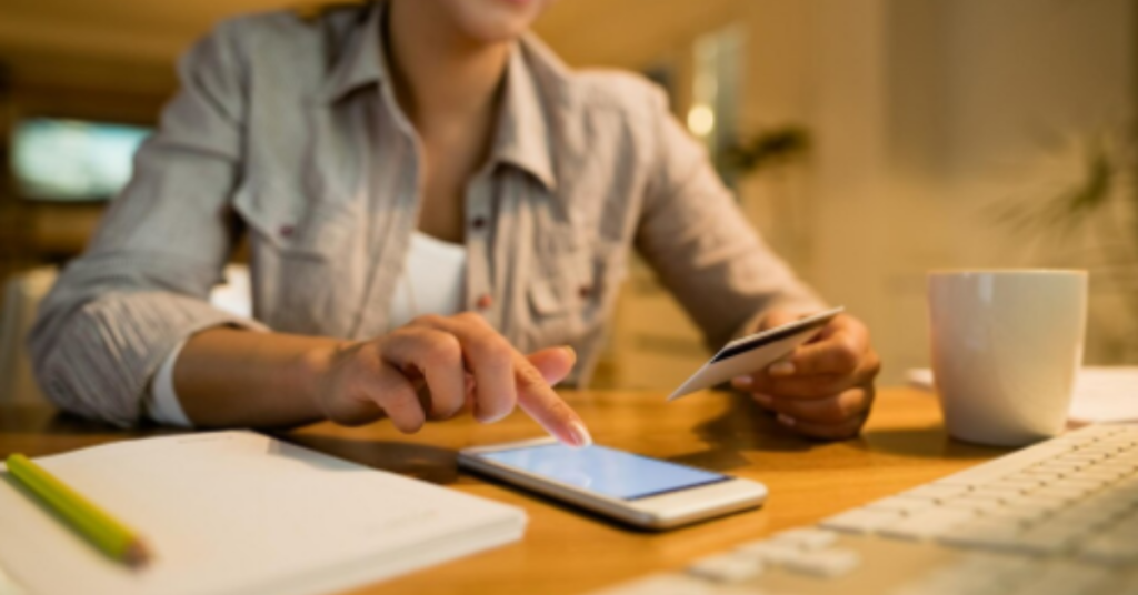 Mulher sorridente usando o celular e segurando um cartão de crédito, fazendo uma compra online em casa.