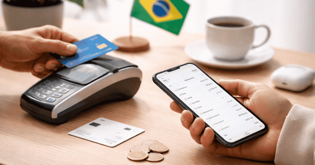 Imagem que representa pagamentos digitais e novas soluções financeiras no Brasil. Sobre uma mesa de madeira, uma pessoa segura um smartphone com uma aplicação financeira aberta que mostra um histórico de transações ou gestão de pagamentos. Ao lado, outra mão aproxima um cartão bancário azul de um terminal de pagamento eletrónico (POS), sugerindo um pagamento por aproximação ou cartão. Na mesa também estão algumas moedas e um cartão adicional pousado, indicando diferentes formas de pagamento. Ao fundo, desfocado, vê-se uma pequena bandeira do Brasil e uma chávena de café, criando um ambiente doméstico ou de escritório. A cena ilustra a utilização moderna de tecnologias financeiras, combinando smartphone e cartão para realizar pagamentos rápidos e acompanhar movimentações financeiras.